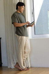 Asian man standing barefoot on floor near window wearing olive tee using tablet, copy space © wavebreak3