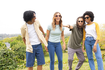 Fototapeta premium Diverse friends laughing, pointing and standing on grassy hilltop wearing sunglasses and wristwatch