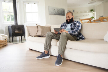 Obraz premium African man sitting on light beige sofa in bright living room holding tablet and smiling