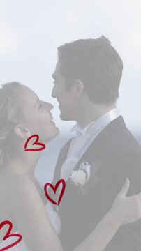 Animation of red hearts over caucasian couple wearing wedding clothes and talking