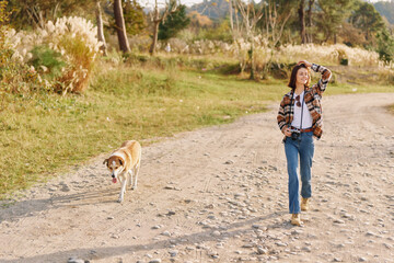 Obraz premium Woman walking her dog along a gravel path in a sunny countryside scene, casual attire, open landscape, and a peaceful outdoor moment of companionship and exercise.