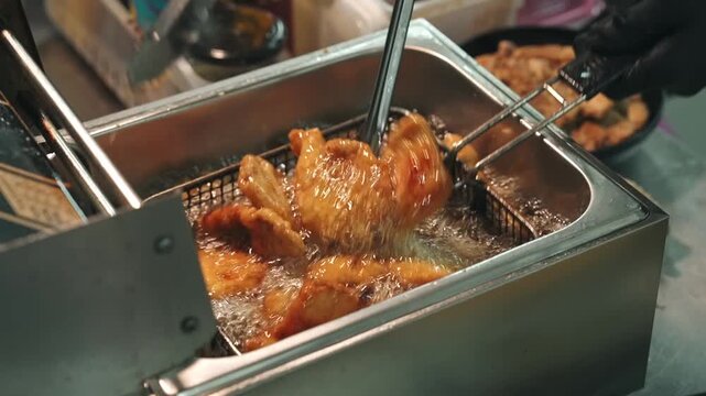 Close-up tracking shot of a chef using tongs to submerge and turn golden battered pieces inside a wire basket submerged in bubbling deep fryer oil.