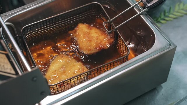 High-angle static shot of seasoned chicken breasts deep frying in bubbling golden oil inside a stainless steel countertop fryer basket.
