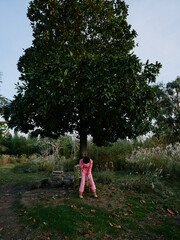 Obraz premium Child in pink clothing stands beneath a large tree in a park, autumn leaves scattered on the ground, conveying exploration and connection with nature.
