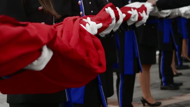 Cinematic close-up shot of an honor guard line wearing black and blue uniforms and white gloves, carefully holding and carrying a long, folded red, white and green Mexican ceremonial flag outdoors.