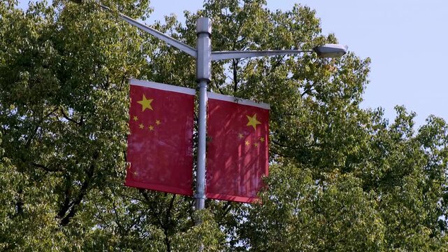 Chinese national flag banners hang displayed on a streetlight post against lush green trees in a city.