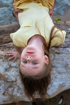 Young girl with blue eyes hanging upside down on a log, smiling playfully, wearing a yellow shirt in a natural outdoor setting.