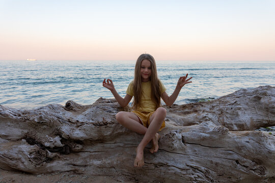 Young girl in yellow dress sits cross-legged on driftwood, meditating by the calm sea at sunset, serene and peaceful atmosphere.