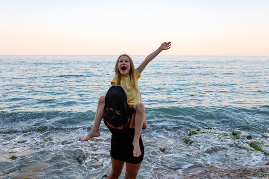 Happy mother lifting her daughter by the sea at sunset, expressing excitement and fun, with waves and clear sky in the background.