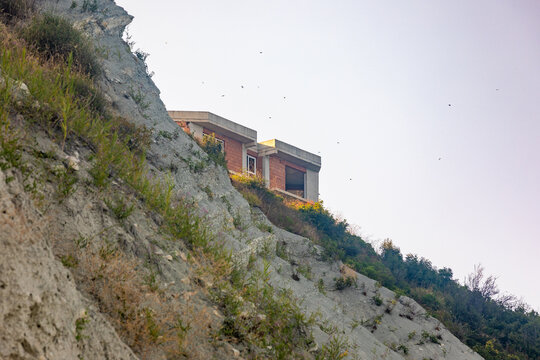 Contemporary house perched on a steep hillside with sparse vegetation and clear sky, offering a unique architectural view in a natural setting.