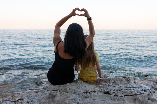 Back view of woman and child sitting on driftwood, making a heart shape with hands, overlooking calm ocean at sunset.