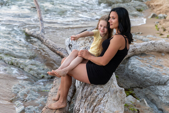 Woman and young girl sit on driftwood by the ocean, enjoying a peaceful moment together on a rocky shoreline.