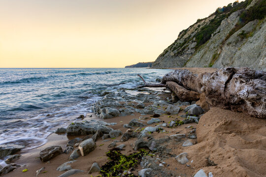 Serene rocky shoreline with gentle waves, driftwood, and a dramatic cliff at sunset, ideal for travel, nature, and wellness concepts.