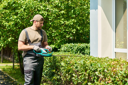 Caucasian male gardener trimming green hedge with electric trimmer near house. Image suitable for professional landscaping services and home maintenance marketing
