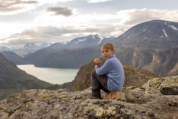 Fototapeta premium Beautiful european family with children and pet dog hiking Knutshoe summit with blue lake in mountain landscape in Jotunheimen National Park in Norway, mountains of Besseggen in background, cloudy sky