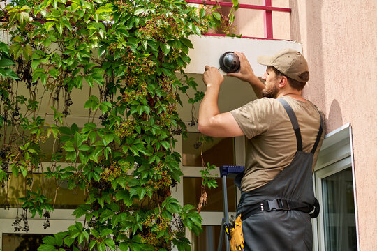Caucasian young adult male technician standing on ladder and installing security camera on building wall. Image represents home safety and is suitable for security service marketing