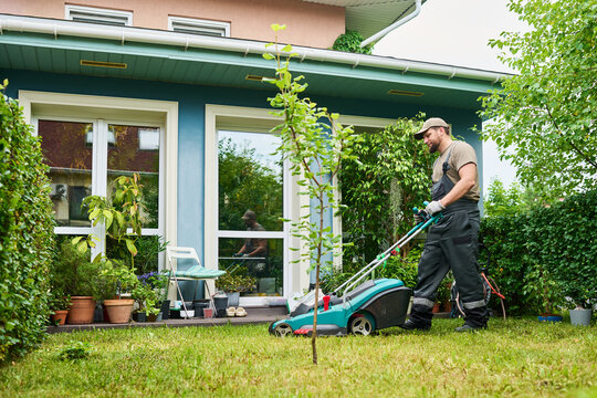 Caucasian young adult male gardener pushing lawn mower on green grass in backyard. Image represents professional landscaping services and residential property maintenance, wide shot