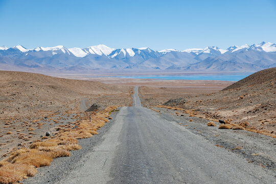 Straight highway road leading down toward alpine blue Karakul Lake and snow peaks in Eastern Pamir, Tajikistan