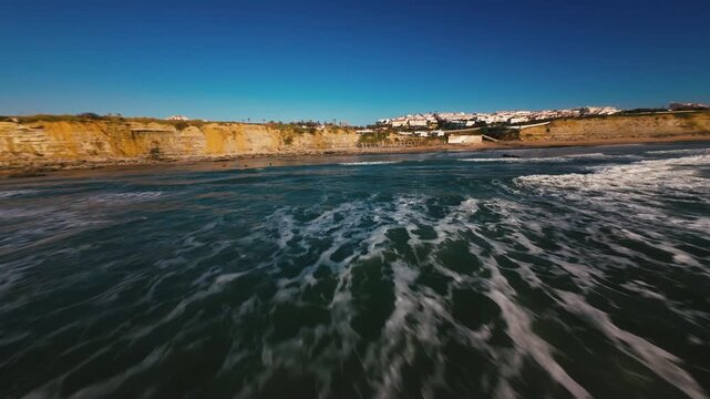 Aerial view of ocean waves crashing against rocky shores. Clear blue sky above calm sea. Coastal cliffs with greenery and distant houses. Perfect for travel, nature, or coastal themes