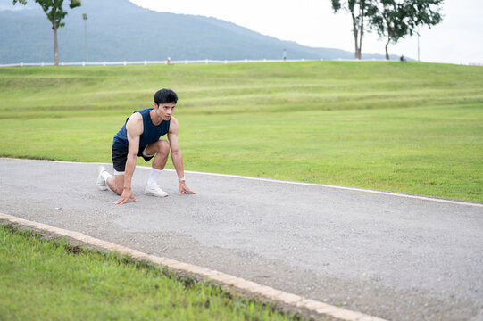 Athletic Asian man sitting crouching preparing to run on the road along a wide green park landscape.