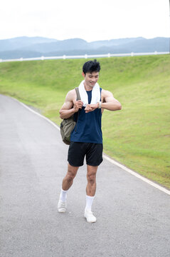 Athletic Asian man checking time on watch while walking on a quiet road surrounded by green hills.