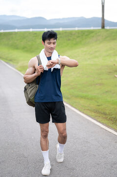Athletic Asian man checking time on watch while walking on a street road surrounded by green hills.