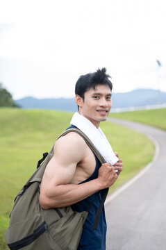 Athletic Asian man carrying a backpack and walking along the road after exercising in greenery park.