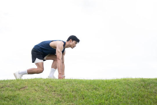 Athletic Asian man crouching preparing to run on the road over a grassy field under clear bright sky