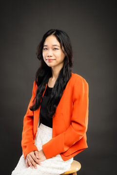 Portrait of a smiling young Southeast Asian Malaysian woman wearing professional corporate attire, featuring a bright orange blazer and black top with a confident pose