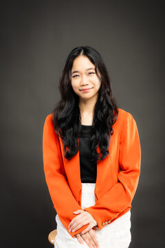 Portrait of a smiling young Southeast Asian Malaysian woman wearing professional corporate attire, featuring a bright orange blazer and black top with a confident pose