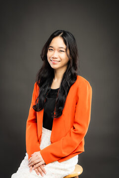Portrait of a smiling young Southeast Asian Malaysian woman wearing professional corporate attire, featuring a bright orange blazer and black top with a confident pose