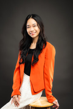 Portrait of a smiling young Southeast Asian Malaysian woman wearing professional corporate attire, featuring a bright orange blazer and black top with a confident pose
