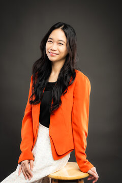Portrait of a smiling young Southeast Asian Malaysian woman wearing professional corporate attire, featuring a bright orange blazer and black top with a confident pose