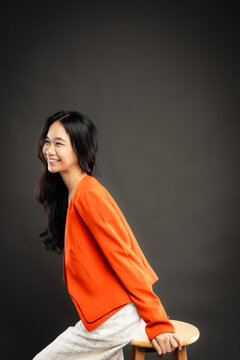 Portrait of a smiling young Southeast Asian Malaysian woman wearing professional corporate attire, featuring a bright orange blazer and black top with a confident pose