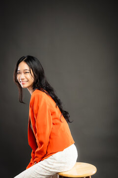 Portrait of a smiling young Southeast Asian Malaysian woman wearing professional corporate attire, featuring a bright orange blazer and black top with a confident pose