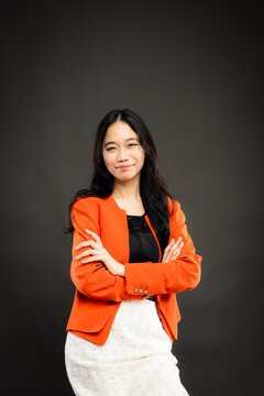 Portrait of a smiling young Southeast Asian Malaysian woman wearing professional corporate attire, featuring a bright orange blazer and black top with a confident pose