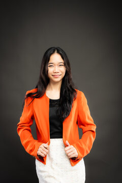 Portrait of a smiling young Southeast Asian Malaysian woman wearing professional corporate attire, featuring a bright orange blazer and black top with a confident pose