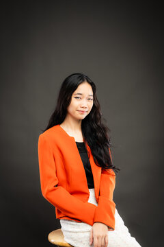 Portrait of a smiling young Southeast Asian Malaysian woman wearing professional corporate attire, featuring a bright orange blazer and black top with a confident pose