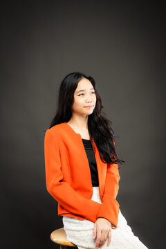 Portrait of a smiling young Southeast Asian Malaysian woman wearing professional corporate attire, featuring a bright orange blazer and black top with a confident pose