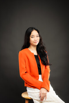 Portrait of a smiling young Southeast Asian Malaysian woman wearing professional corporate attire, featuring a bright orange blazer and black top with a confident pose