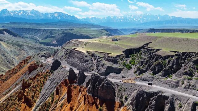 Aerial view of a flock of sheep and a vehicle on a winding dirt road along the edge of a deep colorful canyon in Anjihai Grand Canyon, Xinjiang, China