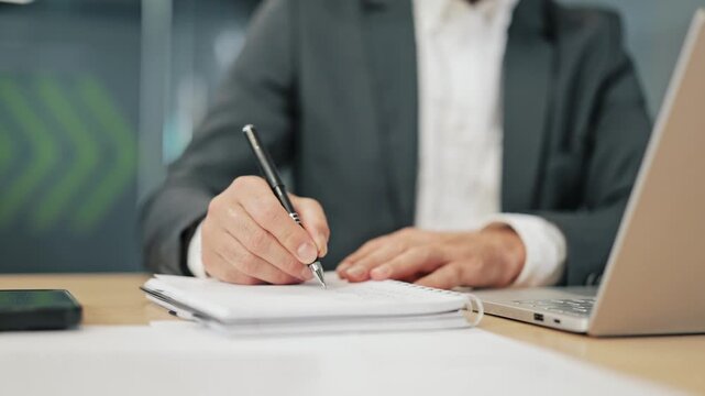 Business person diligently takes notes on paper in office setting. Work attentively at wooden desk with laptop, smartphone. Concentrated on task at hand, important information for project planning.