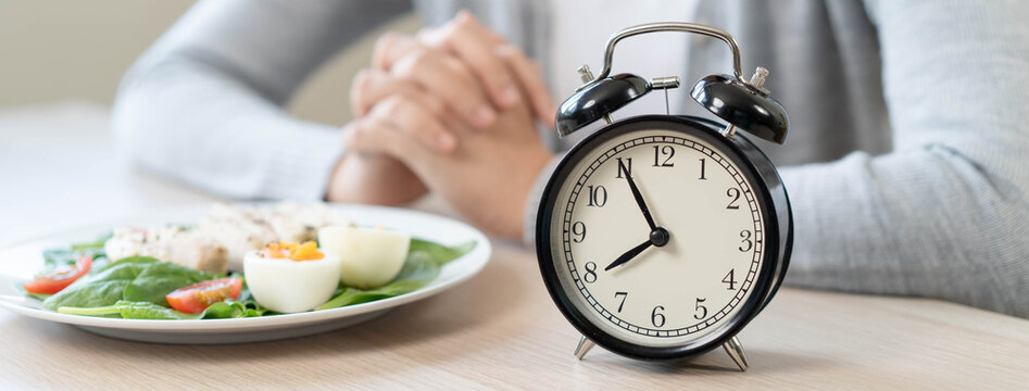 Woman waiting to eat her healthy meal during intermittent fasting concept with alarm clock on wooden table background