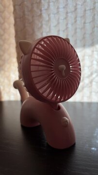 Pink portable handheld mini fan with cute cat ear design cooling air during a summer heatwave, providing a refreshing breeze from its spinning blades at home