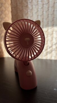 Portable pink mini fan with cat ears rotating on a table for cooling air, small electric battery powered blower ventilator for freshness and hot weather relief