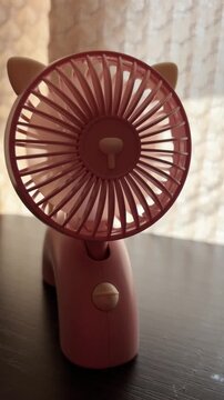 Pink portable handheld mini fan with cat ears design spinning its blades on a dark table, creating a gentle breeze on a warm and sunny summer day in a modern room