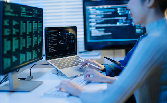 Two female programmers collaborate late in a modern tech office, analyzing computer code on screens to optimize software development
