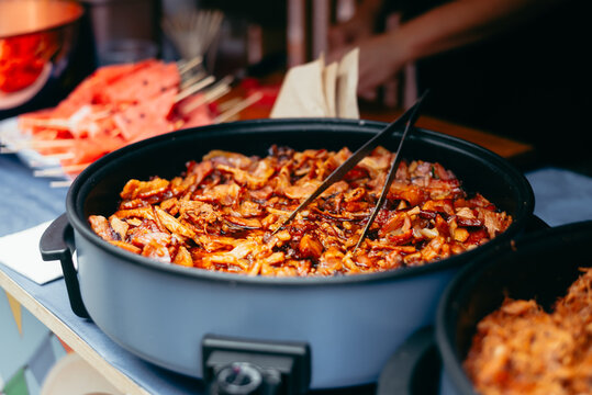 A large serving dish holds a baked food item, placed on a table at an outdoor event. Nearby, skewers of watermelon and other dishes add to the array of food offerings