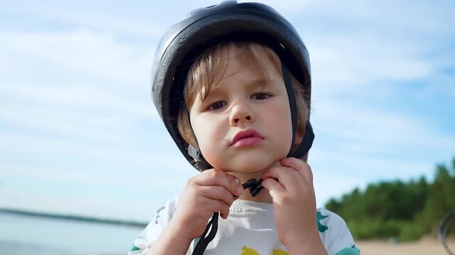 A focused toddler secures a black helmet, adjusting straps while preparing for a bike ride. This determined child ensures safety first against falls under a cloudy sky.