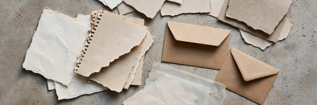 Tactile flat lay of various blank paper sheets with ripped notebook edges and envelopes on matte beige concrete